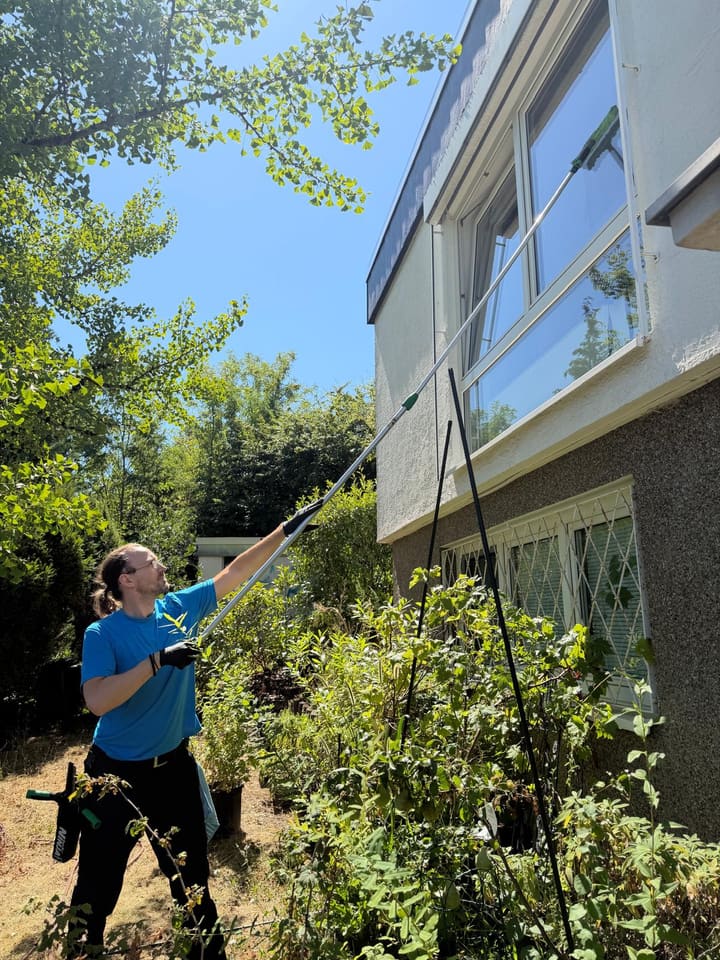 Mitarbeiter reinigt ein Obergeschossfenster mit der Teleskopstange vom Garten aus